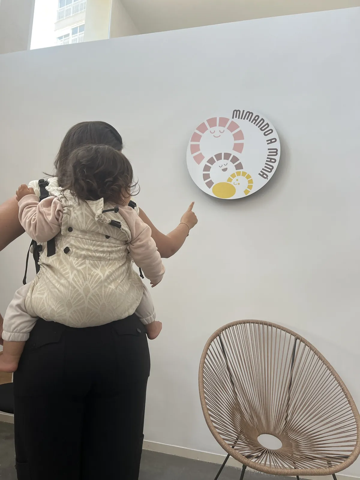 Asesoría de porteo en Alicante: niña en mochila ergonómica señalando el logo de Mimando a Mamá