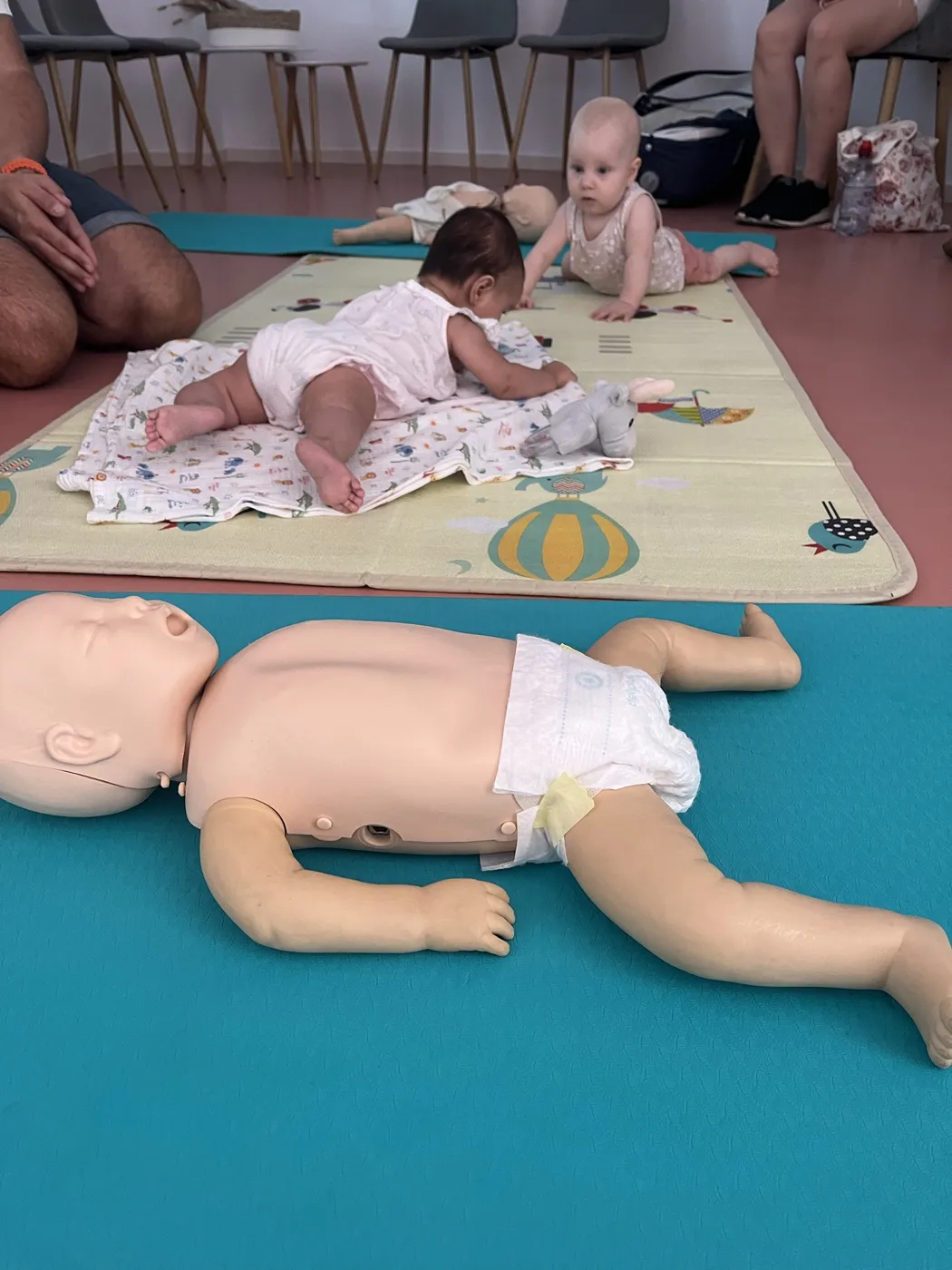 Bebés jugando en la alfombra junto a un maniquí de RCP durante el taller babyfriendly de primeros auxilios en Alicante